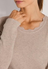 Frau in einem beigen ribbelten Pullover mit gewelltem Ausschnitt, die ihre Hand nah am Hals hält.