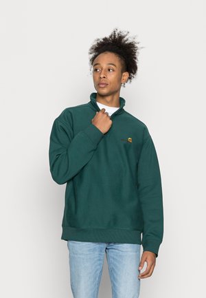 Sweatshirt verde escuro com gola alta e punhos canelados. Apresenta um pequeno logótipo no peito. Usado com jeans azul claro.