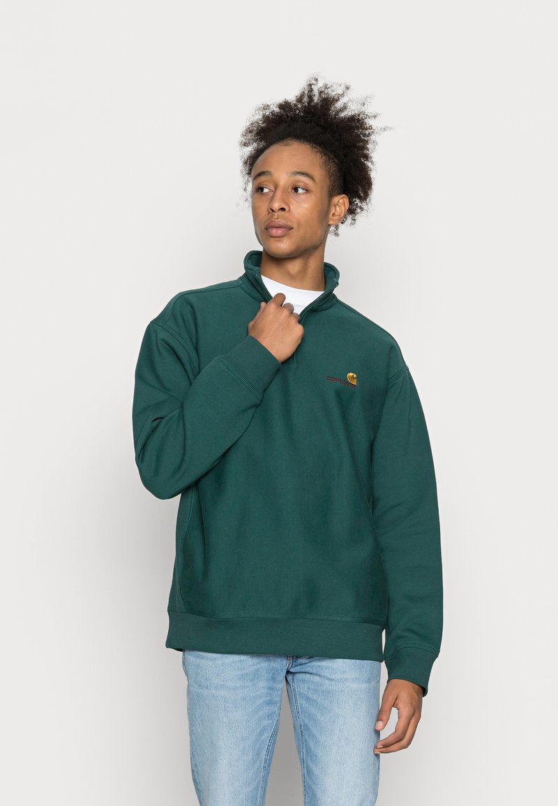 Sweatshirt verde escuro com gola alta e punhos canelados. Apresenta um pequeno logótipo no peito. Usado com jeans azul claro.