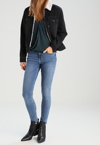 Schwarze Jeansjacke mit weißem Kunstfellkragen, kombiniert mit einem türkisfarbenen Oberteil und hellblauen Skinny-Jeans. Schwarze Stiefeletten runden den Look ab.