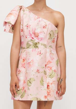 Robe rose à une épaule avec motif floral et grand nœud sur l'épaule, portée par une personne avec tatouages visibles sur les bras et peau claire.