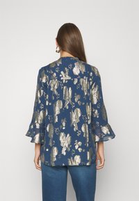 Blusa azul marino con patrón floral plateado, mangas acampanadas de tres cuartos y un corte relaxed. Tejido ligero con una textura suave.