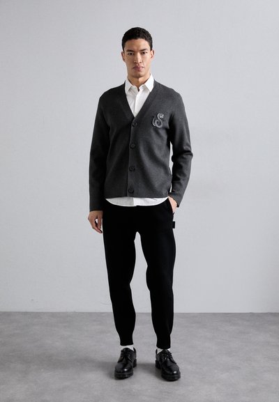 Cardigan grigio antracite con scollatura a V e cinque bottoni, caratterizzato da un dettaglio ricamato decorativo sul petto, abbinato a pantaloni neri e scarpe.