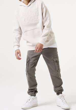 Person trägt einen cremefarbenen Hoodie mit erhabenem Schriftzug, graue Cargohosen und weiße Sneaker, steht mit angewinkelten Knien und entspannten Händen.