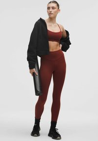 Sportos szett fekete crop púlóverrel, bordó sportmelltartóval és hozzá illő magas derekú leggingssel. Fekete jóga matot tart.