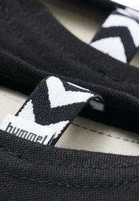 Sneaker textil negru cu etichetă albă în model chevron și detalii de cusături. Prim-plan al materialului și elementelor de design.