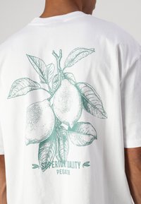 Biela tričko sľubujem svetlozelenou ilustráciou citrónov a listov na chrbte, s textom "SUPERIOR QUALITY PEGADOR."