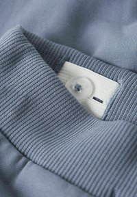 Hellblaues Gewebe mit gerippter Textur, ausgestattet mit einem weißen gestickten Etikett und einem klaren Knopf. Nahaufnahme des Textildetails.