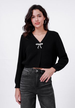 Pull court noir avec texture côtelée, décolleté en V et détail de nœud en perles au centre. Assorti avec un jean taille haute gris.