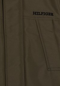 Giacca verde oliva con una texture liscia, caratterizzata da un prominente logo "HILFIGER" ricamato in nero. Visibili molte tasche e dettagli cuciti.