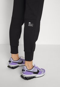 Svarte joggebukser med Lacoste-logo detalj, paret med retro-inspirerte sneakers med lilla og svarte aksenter, hvite lisser og synlig demping.