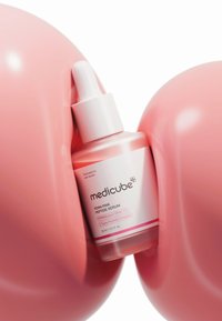 Siero Medicube Peptide in una bottiglia di vetro rosa rotonda con un tappo contagocce, etichettato "PORN PINK", circondato da due sfere rosa lucide.