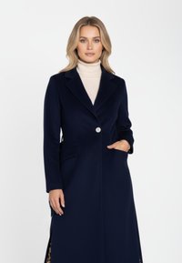 Marineblauer, taillierter Mantel mit einem einzelnen Knopf, Revers mit Kerbkragen und seitlichen Taschen; zeichnet sich durch eine glatte Textur und eine lange Silhouette aus.