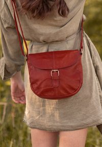 Červená kožená crossbody taška s kulatým tvarem, klopovým uzavřením a detailem přezky. Popruh je nastavitelný a skvěle se hodí k neformálnímu outfitu.
