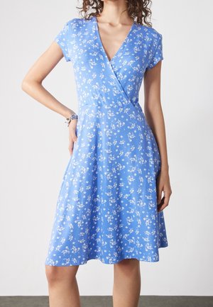 Frau trägt ein blaues, knielanges Wickelkleid mit Blumenmuster und kurzen Ärmeln, steht mit einer Hand auf der Hüfte vor einem einfarbigen Hintergrund.