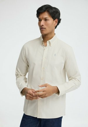 Man met donker haar draagt een beige overhemd met knopen en lange mouwen en staat tegen een effen witte achtergrond.