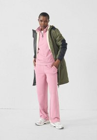 Roze fleece pullover en broek, gecombineerd met een olijfgroene en zwarte jas. De outfit heeft een rits en zilveren sneakers.
