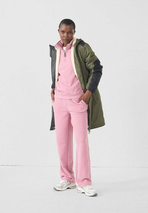 Pull en polaire rose et pantalon assorti, accompagnés d'une veste verte olive et noire. La tenue comprend une fermeture éclair et des baskets argentées.