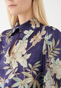 Camicia floreale con sfondo viola decorato da fiori azzurri e beige, colletto e un dettaglio a taglio vicino al collo.