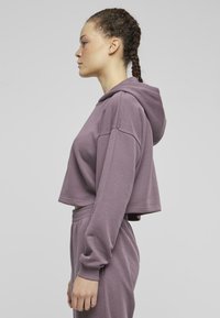 Cropped hoodie viola con maniche lunghe e cappuccio, caratterizzato da una texture morbida. Abbinato a pantaloni jogger coordinati, mostrato di lato.