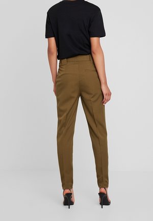 Person trägt olivgrüne eng anliegende Hosen, ein schwarzes kurzärmeliges Top und schwarze Sandalen mit hohen Absätzen, steht vor einem schlichten hellen Hintergrund.