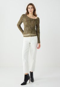Haut à manches longues dans un motif abstrait marron et jaune, avec un décolleté large et une coupe ajustée, associé à un pantalon blanc à jambes droites et des bottes noires.