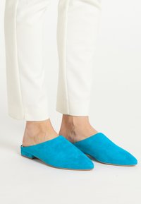 Muli din piele albastră cu vârful ascuțit și tocul plat, prezentând un design fără spate. Asortate cu pantaloni albi adaptați.