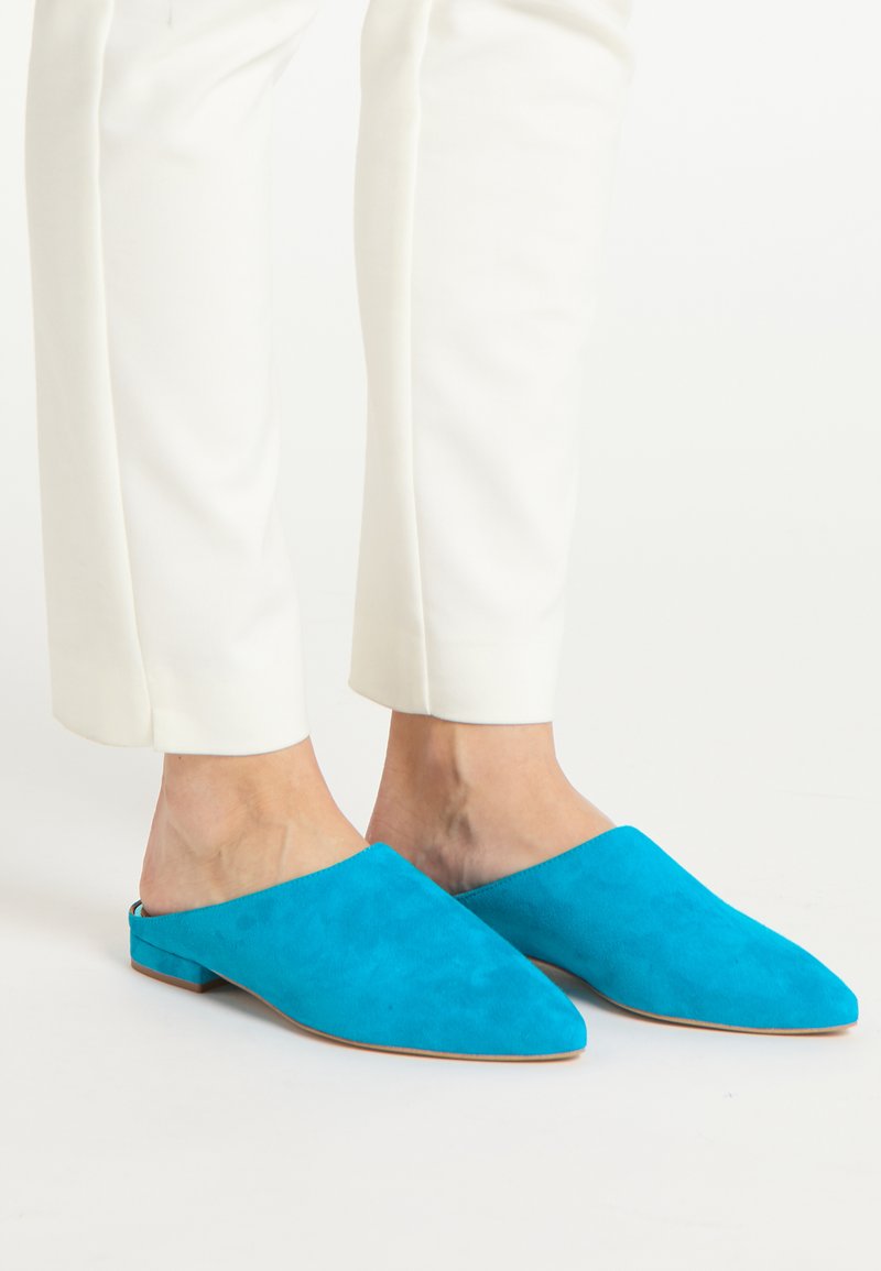 Muli din piele albastră cu vârful ascuțit și tocul plat, prezentând un design fără spate. Asortate cu pantaloni albi adaptați.