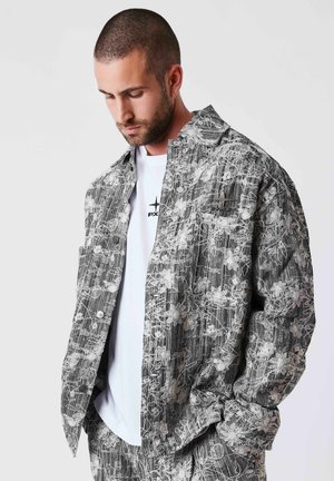 Homme aux cheveux courts et barbe portant une veste grise à motif floral et un pantalon assorti, sur un t-shirt blanc à motif graphique, regardant vers le bas.