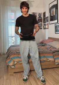 Juodas marškinėliai, šviesiai mėlyni plačiakelniai džinsai, laiko juodą daiktą. Fone matyti lova su raštuota patalynė ir rėminiai plakatai ant sienos.