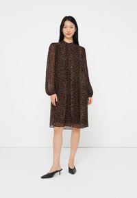 Sieviete stāv, ģērbusies brūnā leopardu raksta garumā līdz ceļiem kleitā ar garām caurspīdīgām piedurknēm un melnās norādītās zemas papēžu kurpēs uz balta fona.