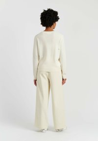 Maglione a maglia color crema con texture a coste e dettaglio del logo sul retro. Abbinato a pantaloni larghi color crema, entrambi in tessuto morbido e leggero.