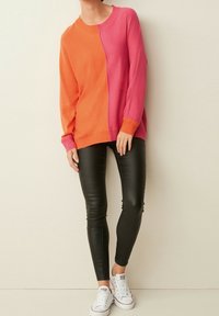 Suéter bicolor en naranja y rosa, con mangas largas, cuello redondo y construcción de punto en capas. Combinado con leggings negros y zapatillas blancas.
