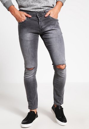 Mężczyzna ubrany w obcisłe szare dżinsy z rozdarciami na kolanach oraz czarne buty typu slip-on, z rękami w kieszeniach, stoi na tle białej ściany.