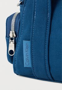 Μπλε υφασμάτινη τσάντα με υφή, διαθέτει ασημένιο φερμουάρ, μικρή ετικέτα με λογότυπο και ορθογώνιο σχήμα που τονίζει τον δομημένο της σχεδιασμό.