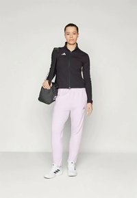 Sieviete melnā aizsietā jakā, gaiši rozā Adidas sporta biksēs, baltos apavos un nesot melnu sporta somu pār plecu.