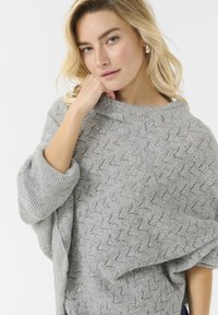 Grå stickad tröja med ett strukturerat zigzag-mönster, vida ärmar och vikbar halsringning. Tyget ser mjukt och löst sittande ut.
