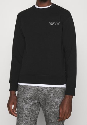 Homme portant un sweat-shirt noir à manches longues avec un petit logo blanc sur la poitrine, un sous-vêtement blanc et un pantalon gris texturé.
