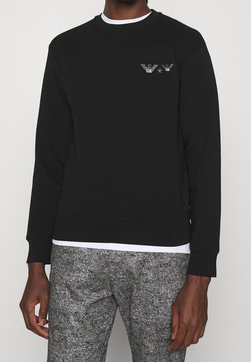 Homme portant un sweat-shirt noir à manches longues avec un petit logo blanc sur la poitrine, un sous-vêtement blanc et un pantalon gris texturé.