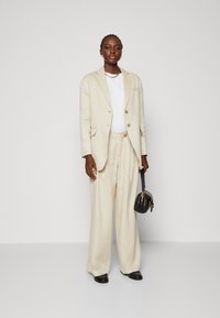 Beige Leinenanzug mit einem taillierten Blazer und weit geschnittenen Hosen. Darunter ein weißes Oberteil, kombiniert mit einer kleinen schwarzen Handtasche.