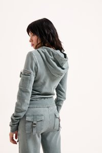 Vrouw met zwart haar, gekleed in een gedempte groene hoodie en bijpassende broek met klepzakken, met de rug naar de camera tegen een effen achtergrond.