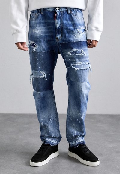 Blue denim jeans with distressed patches, faded texture, and frayed edges. Paired with black shoes featuring white soles.