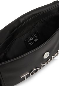 Interior de un bolso sintético negro con una textura suave. Presenta una etiqueta "TOMMY JEANS" y acentos de hardware en plata.