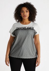 Camiseta gris de manga corta con un patrón de lunares negros, cuello redondo y el texto "DAYDREAMER" en negrita negra en la parte delantera.