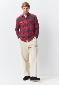 Cămașă flanel în carouri roșii și negre, cu două buzunare la piept. Asortată cu pantaloni bej și pantofi slip-on închiși la culoare, cu talpă texturată.