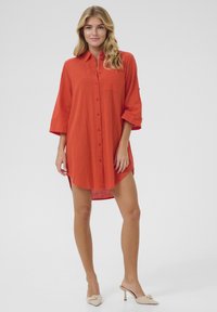 Robe chemise rouge à boutons, avec un col, des manches trois-quarts et des fentes latérales, fabriquée en tissu texturé. Dispose d'une poche poitrine.