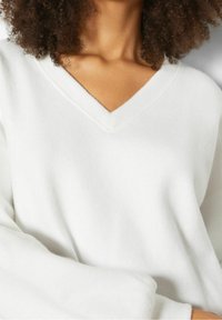 Maglione bianco a V con maniche lunghe, texture morbida e vestibilità comoda. I dettagli delle cuciture sono visibili sul collo e sui polsini.