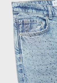 Zone de poche de jean en denim bleu clair avec de petits clous argentés dispersés sur le tissu et des détails de couture visibles.