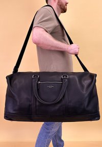 Sort læder duffelbag med tekstureret overflade, dobbelte top hændtag, justerbar skulderrem og en frontlomme med lynlås.