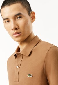 Brun polotshirt gjord av texturerat tyg, med en placket med två knappar och en broderad logotyp på vänster bröst.